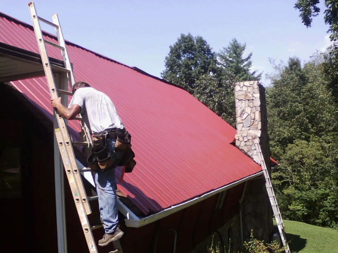 Roofer on a ladder beside a red metal roof and stone chimney — Soffit & Fascia Repair by Roofing Brothers in Sapelo Island